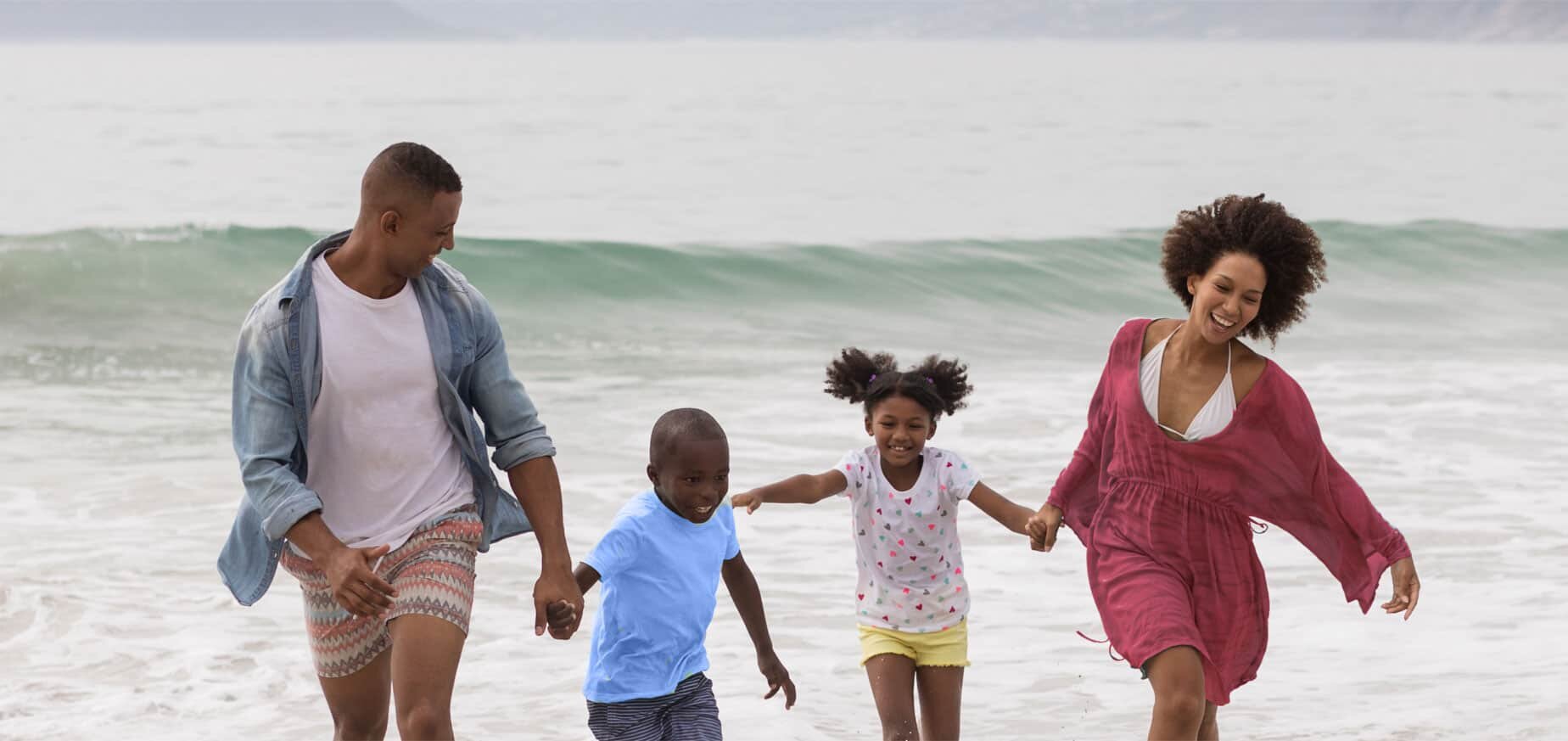 A husband and wife holding hands with their son and daughter. They are standing in shallow water by the beach.
